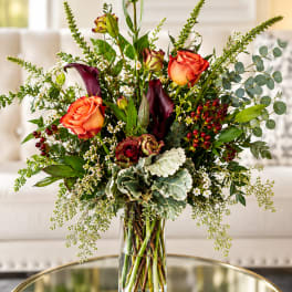 Tall bouquet of orange roses, dark calla lilies, and greenery in a glass vase