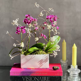 Pink orchid arrangement in a white rectangular planter beside two yellow candles