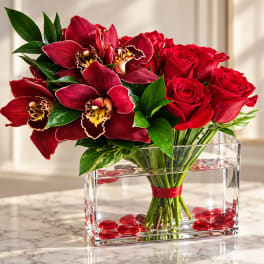 Red roses and burgundy orchids arranged in a clear rectangular vase