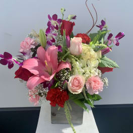 Mixed bouquet with pink lilies, roses, orchids, and carnations in a square vase