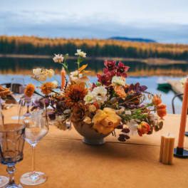 Low centerpiece of yellow and orange flowers in a white bowl on a table by a lake in autumn.