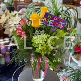Bouquet of yellow roses, pink alstroemeria, and green hydrangea in a glass vase with a birthday sign.