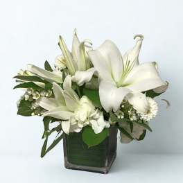 White lilies arranged in a square glass vase with small white accent flowers