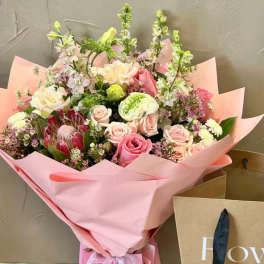 Large pink wrapped bouquet with pastel roses, protea, and mixed flowers beside a kraft shopping bag