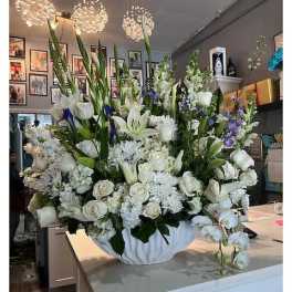 Large white floral arrangement with lilies, roses, and orchids in a white bowl with touches of blue flowers.