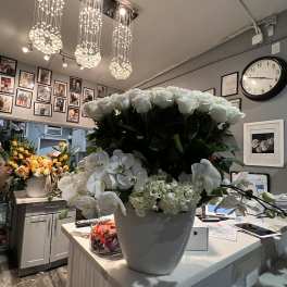 Large white arrangement of roses, orchids, and hydrangea in a white pot on a florist counter