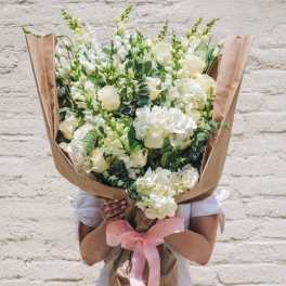 Oversized bouquet of white and cream roses, hydrangeas, and snapdragons wrapped in kraft paper with a pink ribbon.