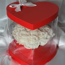 White roses in a red heart-shaped gift box with a clear cover and white ribbon