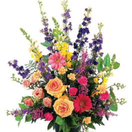 Tall mixed arrangement with peach roses, pink gerbera, yellow flowers, and purple delphinium in a black container.