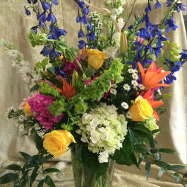 Tall mixed bouquet in a clear glass vase with blue, yellow, orange, and white flowers