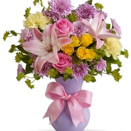 Pink and yellow flower arrangement in a lavender vase with a pink ribbon