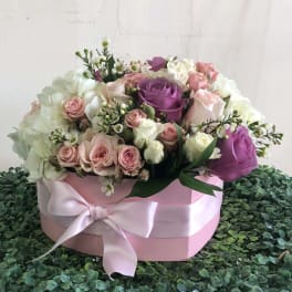 Round pink hatbox of white, blush, and lavender roses with a satin bow on a green textured surface