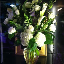 Tall arrangement of white roses, calla lilies, and hydrangeas in a clear glass vase with touches of purple.