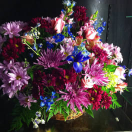 Mixed basket arrangement with pink roses, purple mums, and blue accent flowers in a wicker basket