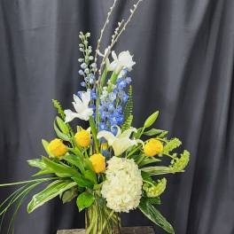 Tall floral arrangement with yellow roses, white lilies, blue flowers, and a white hydrangea in a glass vase
