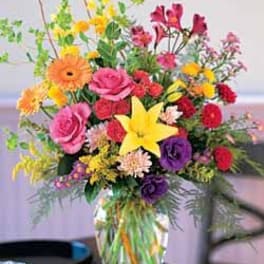 Tall mixed bouquet of pink roses, yellow lily, gerberas, and other bright blooms in a clear glass vase