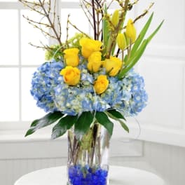 Tall arrangement of yellow roses and tulips with blue hydrangeas in a clear glass vase