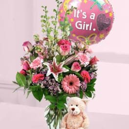 Pink flower arrangement in a glass vase with It’s a Girl balloon and teddy bear.