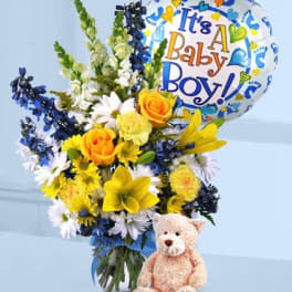 Yellow and white flower bouquet with blue accents, baby boy balloon, and teddy bear in front.