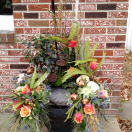 Outdoor arrangement of orange roses, succulents, and pods in a black urn against a brick wall.
