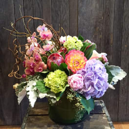 Colorful floral arrangement in a dark green vase with orchids, hydrangeas, and a protea