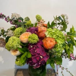Mixed bouquet of orange roses, green hydrangeas, and purple blooms in a glass vase