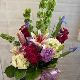 Tall mixed floral arrangement with lilies, hydrangeas, and Bells of Ireland in a glass vase with a lavender bow.