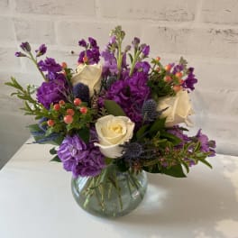 Round glass vase of white roses, purple flowers, blue thistles, and orange berries.