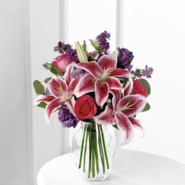 Pink lilies, red roses, and purple flowers arranged in a clear glass vase