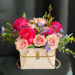 Pink roses and purple daisies arranged in a white handbag-shaped box