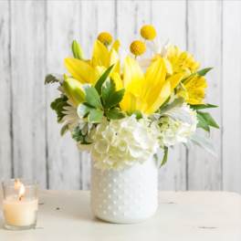 Yellow and white flower arrangement in a white vase beside a lit candle