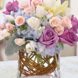 Pastel bouquet of roses, tulips, lilies and hydrangea in a square glass vase with woven branches.