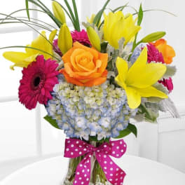 Bright mixed bouquet with yellow lilies, orange rose, pink gerberas, and blue hydrangea in a glass vase.