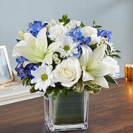 White lilies, roses, daisies, and blue flowers in a square glass vase