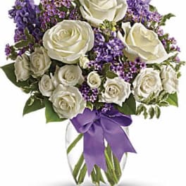 White roses and purple flowers in a clear glass vase with a purple ribbon
