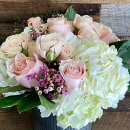 Short arrangement of pale pink roses, white hydrangeas, and small purple blooms in a gray vase