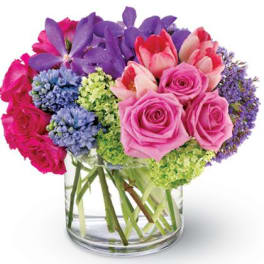 Low glass vase of pink roses, tulips, purple orchids, and blue flowers in a compact arrangement