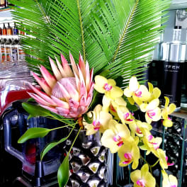 Tropical arrangement with pink protea and yellow orchids in a metallic pineapple vase