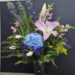 Tall mixed bouquet with pink lilies, blue hydrangea, and purple flowers in a black vase