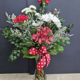 Bouquet of red carnations, white daisies, and pink alstroemeria in a glass vase with a red polka-dot ribbon.