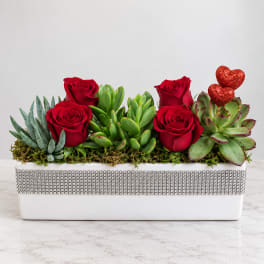 Red roses and succulents arranged in a white rectangular planter with glitter hearts