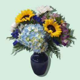 Mixed bouquet with blue hydrangea, yellow sunflowers, white daisies, and purple blooms in a dark blue vase.