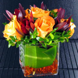 Orange and yellow roses in a clear square vase with green foliage