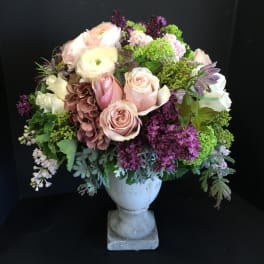 Pastel bouquet of roses, ranunculus, and purple blooms in a white urn vase