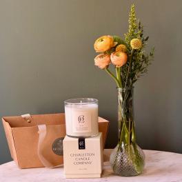 Peach and yellow flower stems in a tall glass vase beside a white candle and tan gift bag