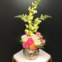 Tall floral arrangement with roses, carnations, and yellow snapdragons in a mirrored vase