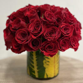 Round arrangement of red roses in a clear glass cylinder vase lined with green and yellow leaves.