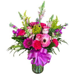 Bright pink and purple mixed flower arrangement in a green glass vase with a large purple bow