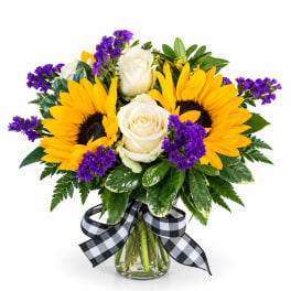 Bouquet of yellow sunflowers, white roses, and purple filler in a glass vase with a checkered ribbon