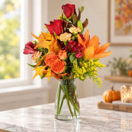 Autumn bouquet of red roses, orange lilies, and yellow filler in a glass vase